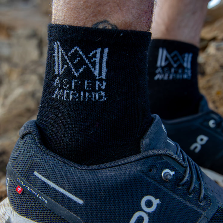 Black socks with 'Aspen Merino' branding worn with blue running shoes on a rocky surface.