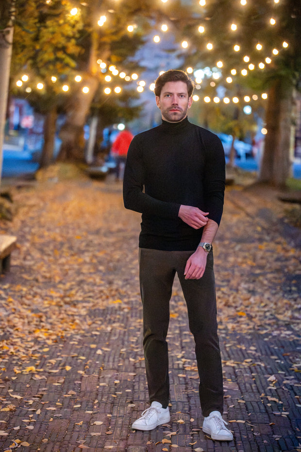 Man in black Merino Turtleneck and dark pants standing on a leaf-covered path with string lights in the background