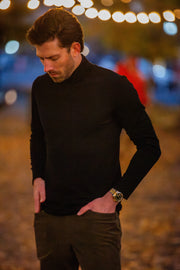 Man wearing a black Merino Turtleneck and brown pants with a blurred background of lights.