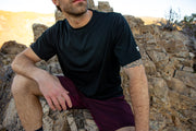 Man sitting on rocky terrain wearing a black Merino t-shirt and maroon shorts.