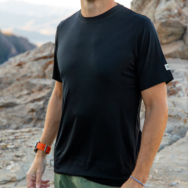 Man wearing a black Merino t-shirt standing on rocky terrain with mountains in the background