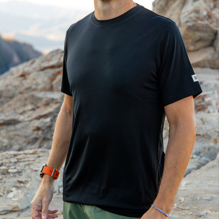 Man wearing a black Merino t-shirt standing on rocky terrain with mountains in the background