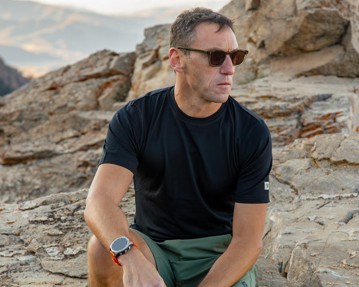 Man sitting on rocky terrain wearing sunglasses and a black Merino shirt.