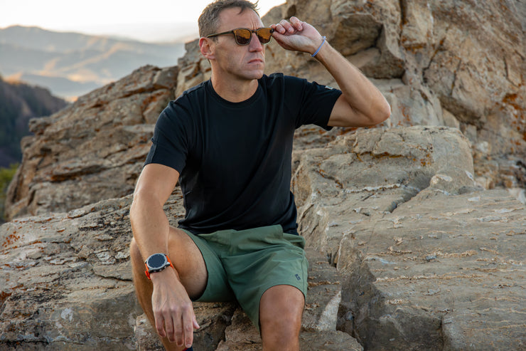 Man sitting on a rocky outcrop wearing sunglasses and a black Merino t-shirt.
